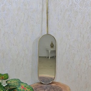 Pode incluir: Um espelho oval dourado com uma moldura longa e fina pendurado em um suporte de teto. O espelho reflete uma sala com uma mesa branca e um objeto decorativo no topo.