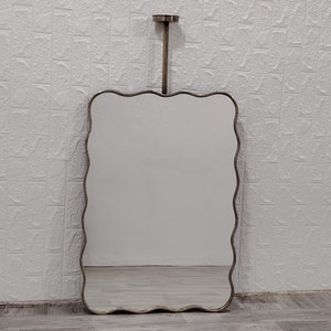 May include: A wavy-edged, rectangular mirror with a silver frame hangs from a silver chain. The mirror is reflecting a white wall with a textured pattern.
