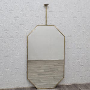 Pode incluir: Um espelho octogonal com moldura de latão e uma haste de latão para pendurar. O espelho reflete a sala. A parede atrás do espelho é branca com um padrão texturizado. O espelho é de cor neutra.