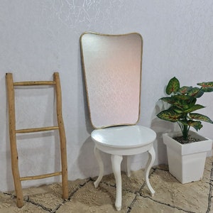 May include: A white round table with four legs and a gold framed mirror with a wavy top. The table and mirror are on a beige rug with a black pattern. A green plant in a white pot is on the right side of the image.