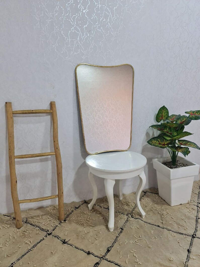 May include: A white round table with four legs and a gold framed mirror with a wavy top. The table and mirror are on a beige rug with a black pattern. A green plant in a white pot is on the right side of the image.