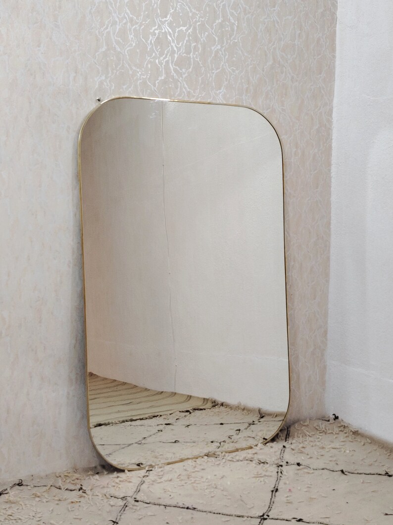 May include: A large, rectangular mirror with a gold-colored frame. The mirror has rounded edges and is leaning against a wall.