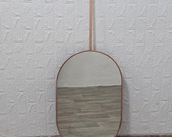 May include: A rose gold oval mirror with a metal frame and a hanging rod. The mirror is hanging on a white brick wall.