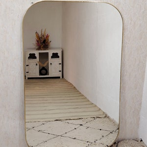 May include: A large, rectangular mirror with a gold-colored frame. The mirror has rounded corners and is reflecting a white cabinet with a vase of dried flowers.
