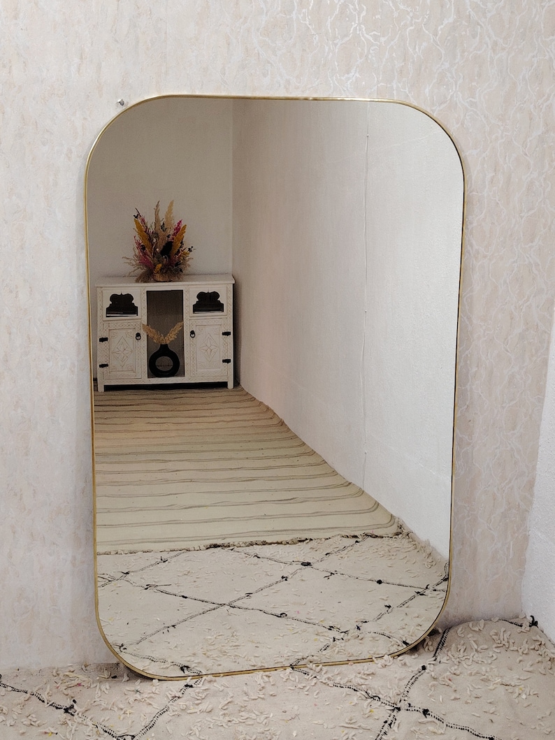May include: A large, rectangular mirror with a gold-colored frame. The mirror has rounded corners and is reflecting a white cabinet with a vase of dried flowers.