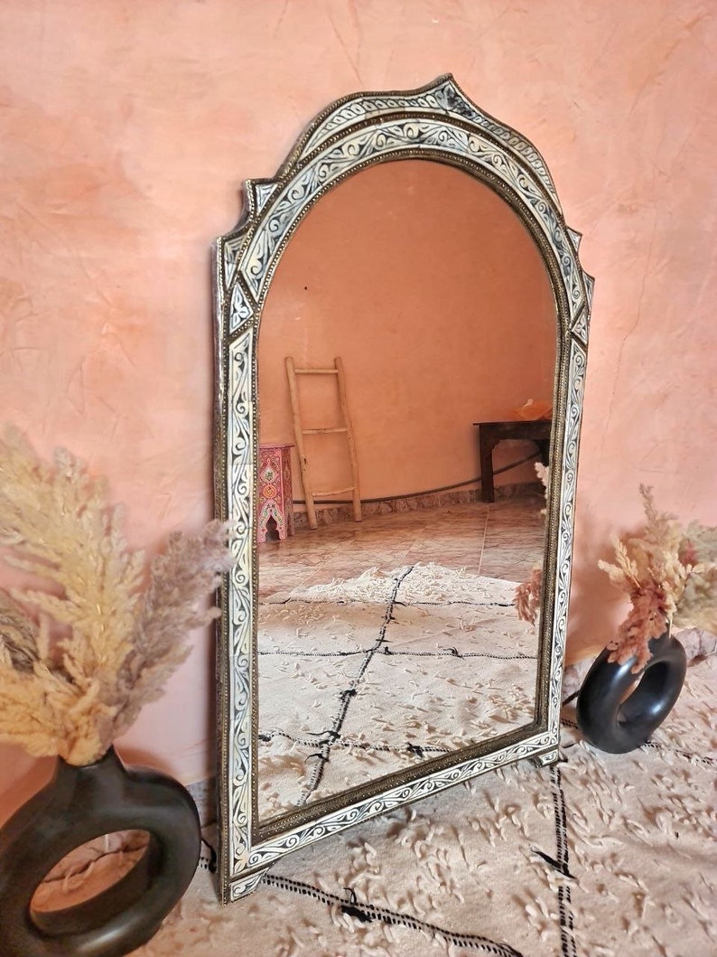 Op de afbeelding: Een grote, sierlijke, gebogen spiegel met een wit en gouden frame. Het frame is versierd met ingewikkelde houtsnijwerk en ontwerpen. De spiegel reflecteert een kamer met een ladder en een houten tafel.