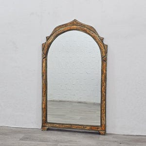 May include: A decorative arched mirror with a carved wooden frame. The frame is painted in a light brown color with gold accents. The mirror is reflecting a white textured wall.