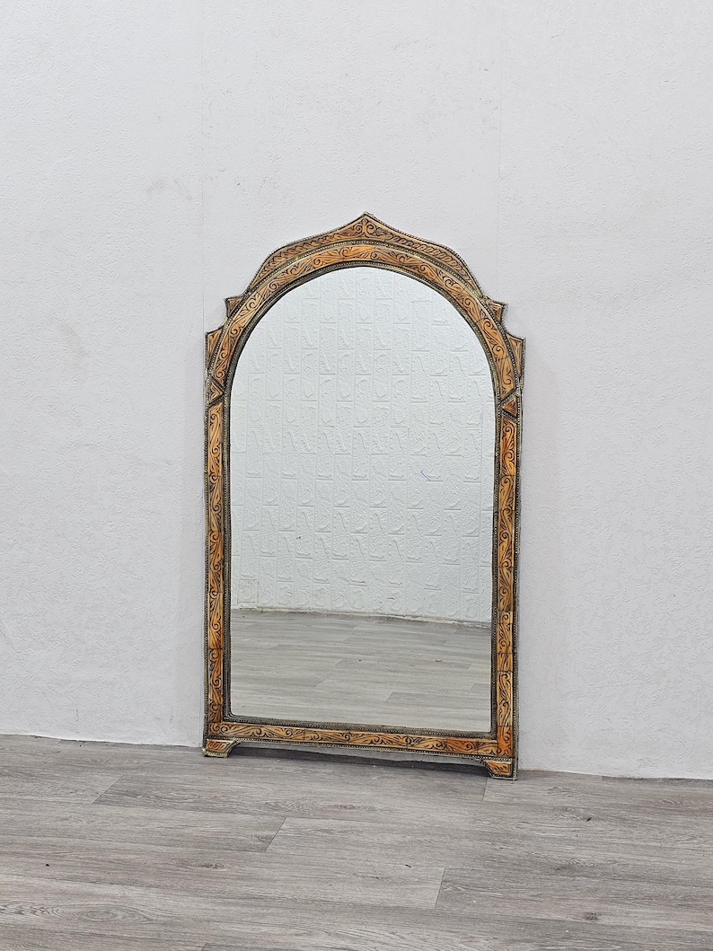 May include: A decorative arched mirror with a carved wooden frame. The frame is painted in a light brown color with gold accents. The mirror is reflecting a white textured wall.