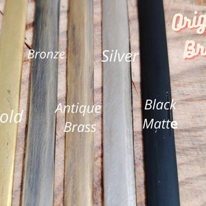 May include: Close-up of five metal strips in different finishes: gold, bronze, antique brass, silver, and black matte. The text "Original Brass!" is written in red on the right side of the image.