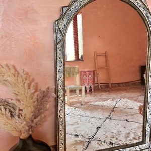 Op de afbeelding: Een grote, sierlijke spiegel met een wit en gouden frame. Het frame is gesneden met ingewikkelde ontwerpen en heeft een licht gebogen bovenkant. De spiegel reflecteert een kamer met een houten ladder en een kleine kruk.