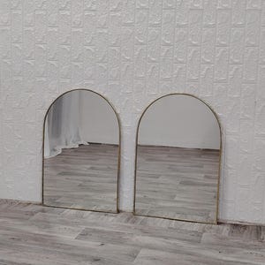 Op de afbeelding: Twee gouden boogspiegels met een witte muur en een houten vloer op de achtergrond.