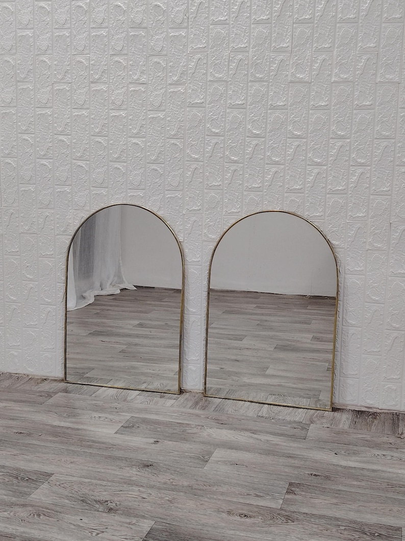 Op de afbeelding: Twee gouden boogspiegels met een witte muur en een houten vloer op de achtergrond.