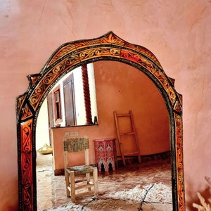 Op de afbeelding: Een decoratieve spiegel met een gesneden houten lijst in een warme, aardse kleurenpalet. De spiegel reflecteert een kamer met een houten stoel, een ladder en een kleine tafel met een kleurrijk ontwerp.