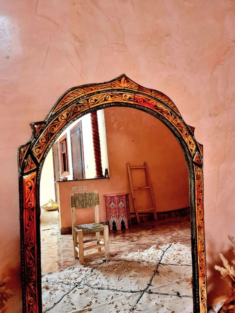 Op de afbeelding: Een decoratieve spiegel met een gesneden houten lijst in een warme, aardse kleurenpalet. De spiegel reflecteert een kamer met een houten stoel, een ladder en een kleine tafel met een kleurrijk ontwerp.