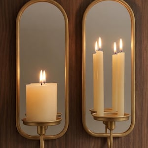 Puede incluir: Dos apliques de pared con marco dorado con espejos ovalados y candelabros. Cada aplique contiene velas encendidas, una con una gruesa vela de pilar y la otra con tres velas delgadas. Los apliques est&aacute;n montados en una pared de madera oscura.