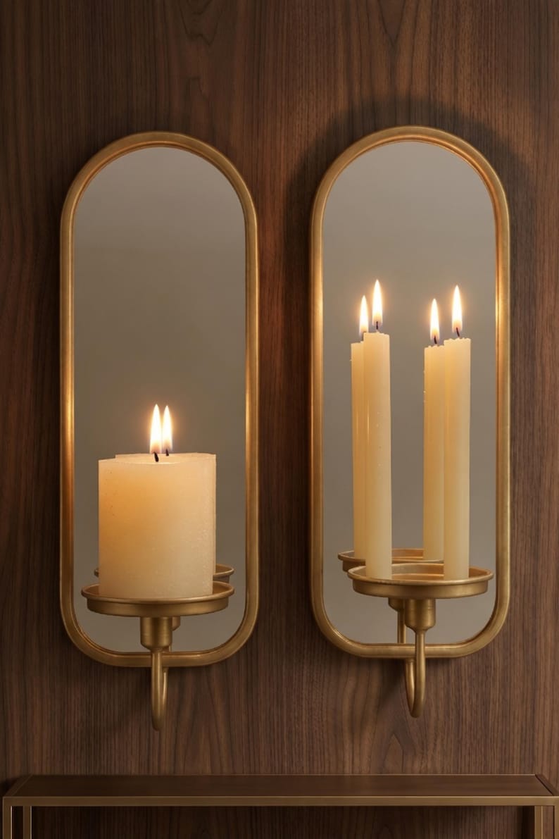 Puede incluir: Dos apliques de pared con marco dorado con espejos ovalados y candelabros. Cada aplique contiene velas encendidas, una con una gruesa vela de pilar y la otra con tres velas delgadas. Los apliques est&aacute;n montados en una pared de madera oscura.