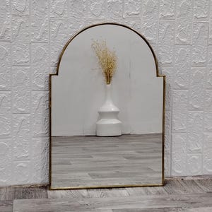 May include: An arched mirror with a gold frame, featuring a white vase with dried flowers inside, reflecting a wooden floor.
