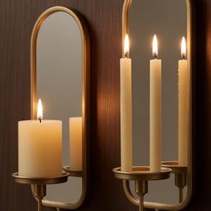 Puede incluir: Dos apliques de pared dorados con espejos y candelabros. Uno sostiene una vela gruesa encendida, el otro tres velas encendidas. Los apliques est&aacute;n montados en una pared de madera marr&oacute;n oscuro, creando un ambiente c&aacute;lido.