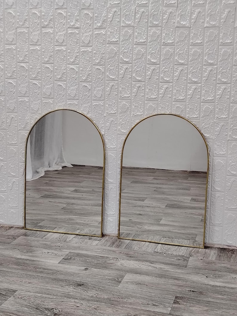 Op de afbeelding: Twee gouden geprofileerde spiegels met een witte achtergrond. De spiegels reflecteren een houten vloer.