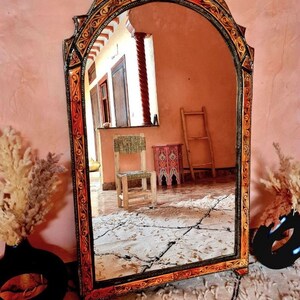 Op de afbeelding: Een grote, sierlijke spiegel met een gesneden houten lijst. De lijst is geschilderd in tinten oranje, goud en zwart. De spiegel reflecteert een kamer met een stoel en een ladder.