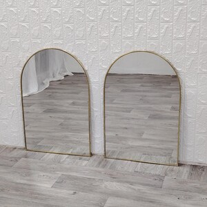 Op de afbeelding: Twee gouden boogspiegels met een reflecterend oppervlak. De spiegels staan op een lichtgrijze houten vloer.