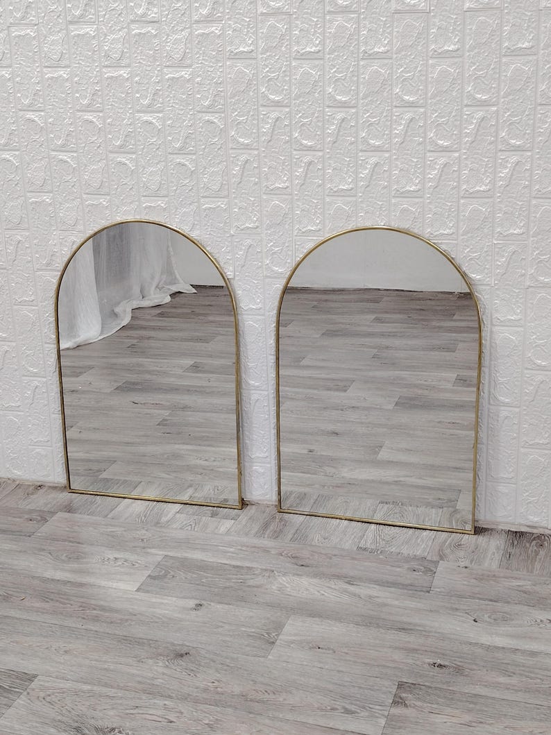 Op de afbeelding: Twee gouden boogspiegels met een reflecterend oppervlak. De spiegels staan op een lichtgrijze houten vloer.