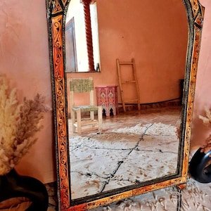 Op de afbeelding: Een grote, sierlijke spiegel met een gesneden houten lijst. De lijst is geschilderd in tinten oranje, geel en zwart. De spiegel reflecteert een kamer met een ladder en een kleine stoel.