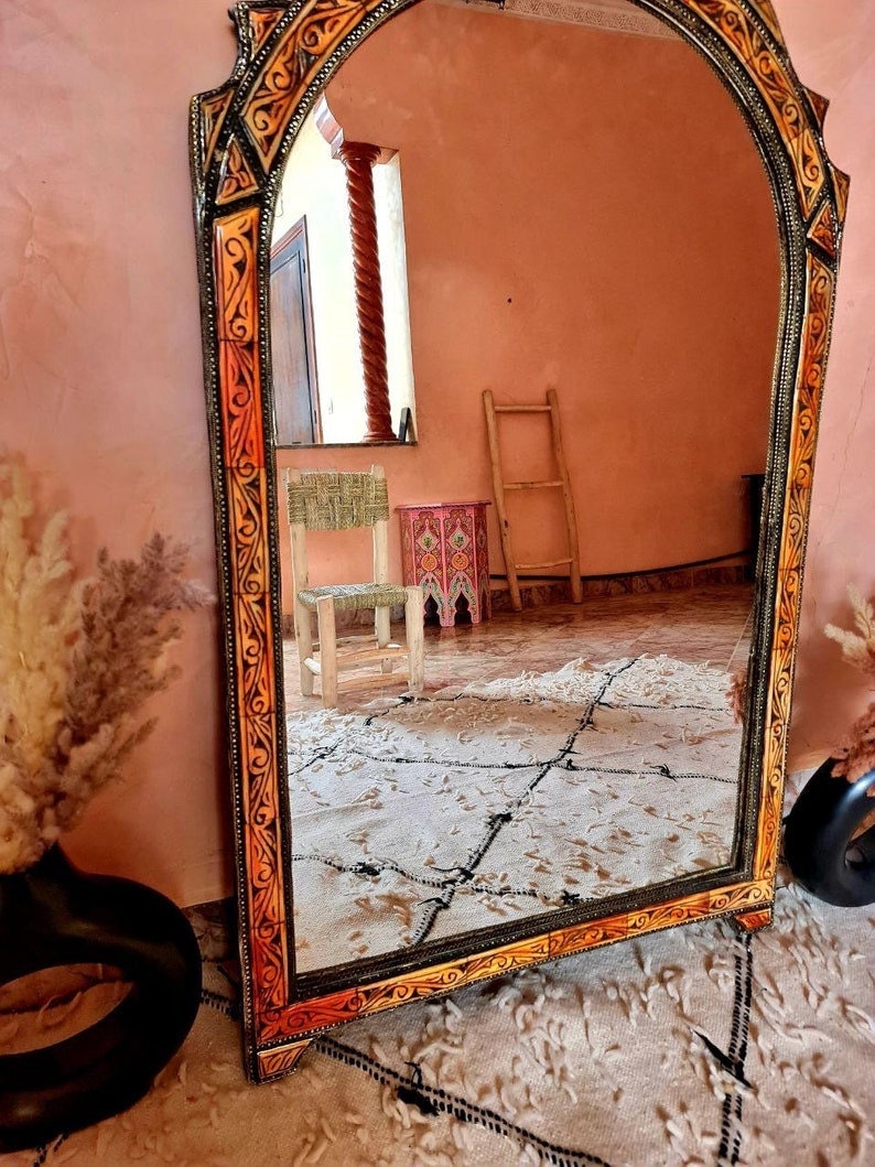 Op de afbeelding: Een grote, sierlijke spiegel met een gesneden houten lijst. De lijst is geschilderd in tinten oranje, geel en zwart. De spiegel reflecteert een kamer met een ladder en een kleine stoel.