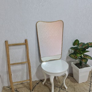 May include: A white vanity table with a round top and three legs. The table has a gold-framed mirror with a wavy top and a white background. A wooden ladder leans against the wall behind the table. A potted plant with green leaves is on the right side of the table.