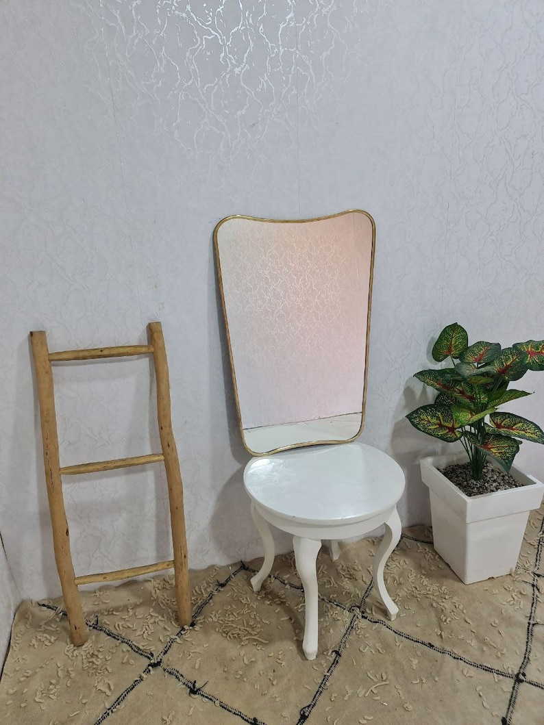 May include: A white vanity table with a round top and three legs. The table has a gold-framed mirror with a wavy top and a white background. A wooden ladder leans against the wall behind the table. A potted plant with green leaves is on the right side of the table.