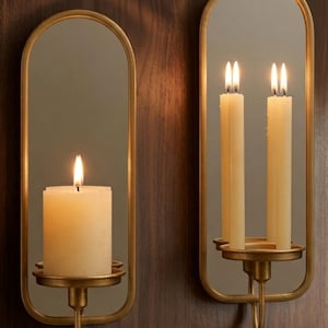 Puede incluir: Dos apliques de pared con marco dorado, espejos y velas encendidas. Uno tiene una vela de pilar gruesa, el otro dos velas delgadas. Los apliques est&aacute;n montados en una pared de paneles de madera oscura, creando un ambiente c&aacute;lido.