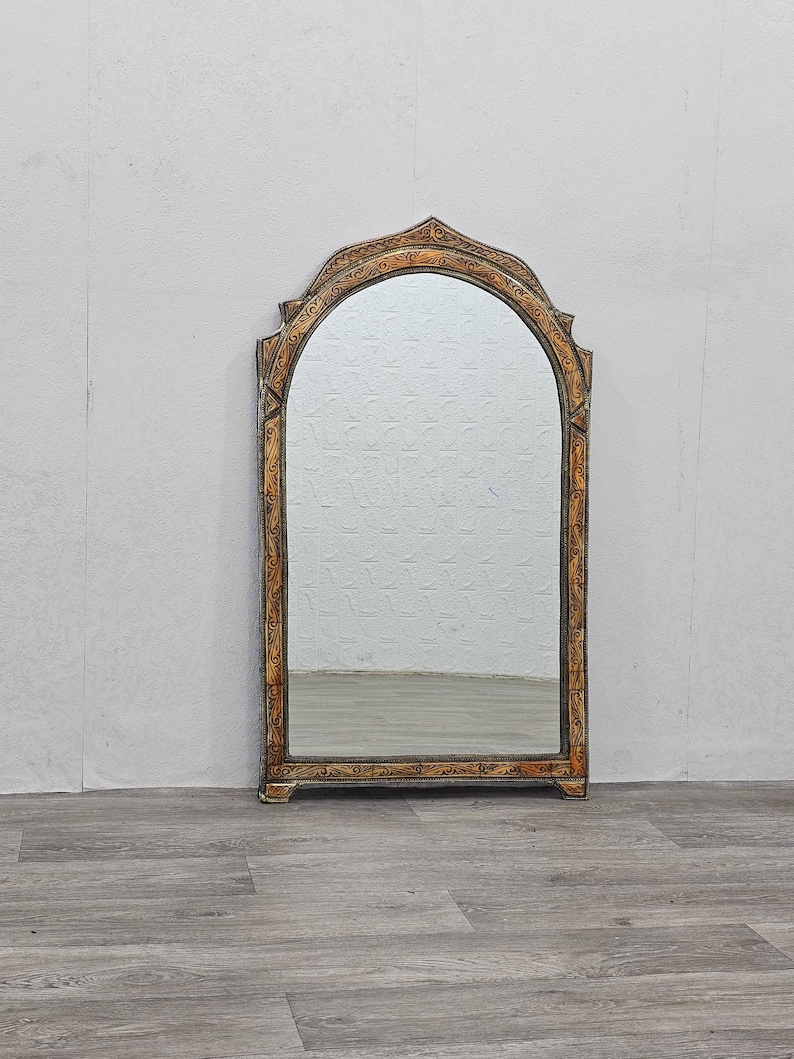 May include: A tall, arched mirror with a decorative, ornate frame. The frame is painted in a gold and brown color scheme with intricate floral designs.