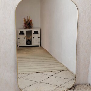 May include: A large, gold-framed, rounded rectangular mirror reflects a white cabinet with a vase of dried flowers. The mirror is leaning against a white wall and is sitting on a white rug with a diamond pattern.