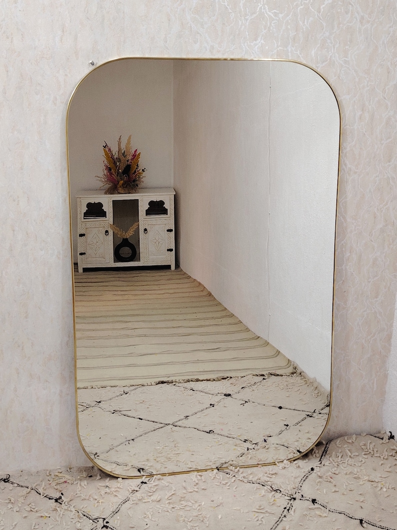 May include: A large, gold-framed, rounded rectangular mirror reflects a white cabinet with a vase of dried flowers. The mirror is leaning against a white wall and is sitting on a white rug with a diamond pattern.