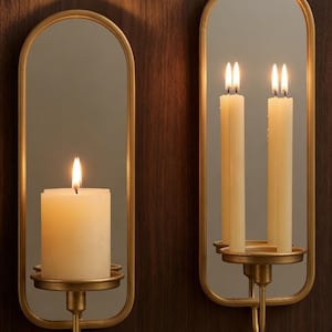 Puede incluir: Dos apliques de pared con marco dorado con espejos ovalados y candelabros. Uno tiene una vela gruesa encendida, el otro dos velas delgadas encendidas. Los apliques est&aacute;n montados en una pared de vetas de madera oscura.
