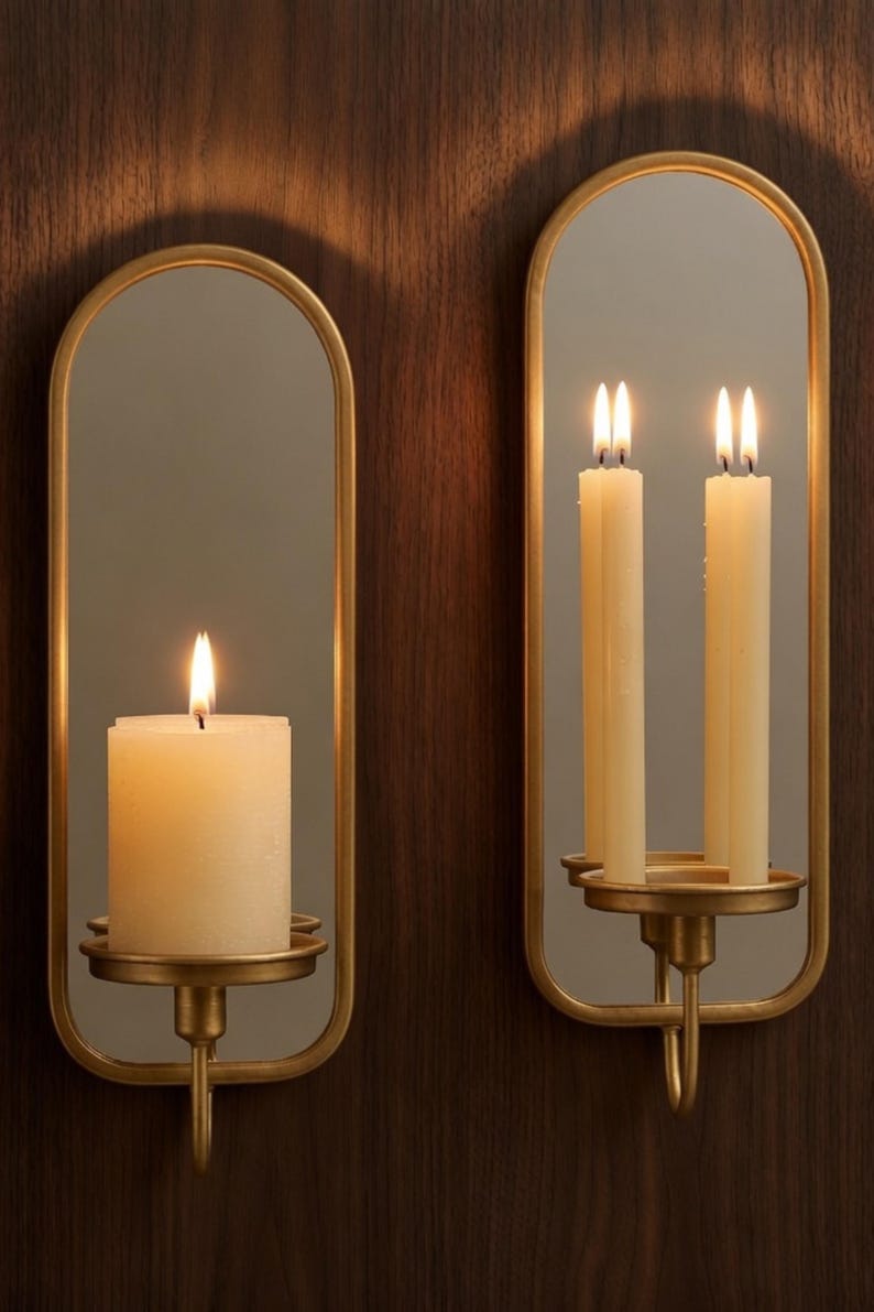 Puede incluir: Dos apliques de pared con marco dorado con espejos ovalados y candelabros. Uno tiene una vela gruesa encendida, el otro dos velas delgadas encendidas. Los apliques est&aacute;n montados en una pared de vetas de madera oscura.
