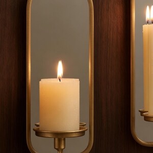 Puede incluir: Una vela de pilar de color marfil encendida en un aplique de pared dorado. El aplique tiene un espejo ovalado y un portavelas decorativo. El fondo es un panel de madera oscura. La llama de la vela es brillante.