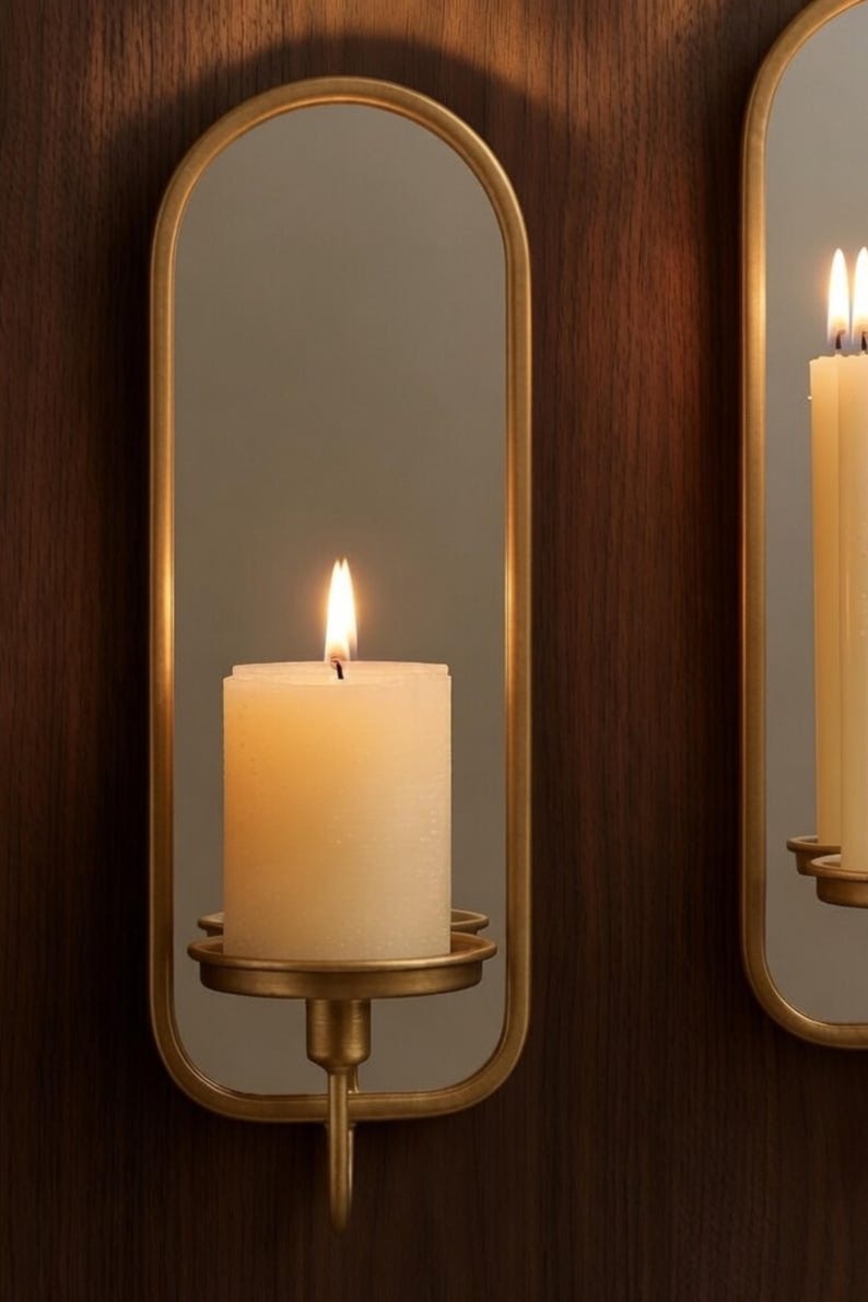 Puede incluir: Una vela de pilar de color marfil encendida en un aplique de pared dorado. El aplique tiene un espejo ovalado y un portavelas decorativo. El fondo es un panel de madera oscura. La llama de la vela es brillante.