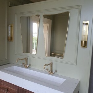 May include: A white bathroom vanity with two gold faucets and a double sink. Two gold framed mirrors hang above the sink. Two gold wall sconces are on either side of the mirrors.