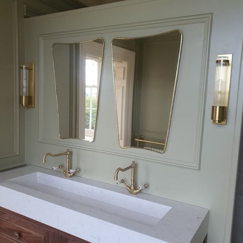 May include: A white bathroom vanity with two gold faucets and a double sink. Two gold framed mirrors hang above the sink. Two gold wall sconces are on either side of the mirrors.
