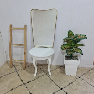 May include: A white vanity table with a round top and three legs. A gold framed mirror with a wavy top sits on the table. A wooden ladder leans against the wall behind the table. A potted green plant with red leaves sits on the floor next to the table.