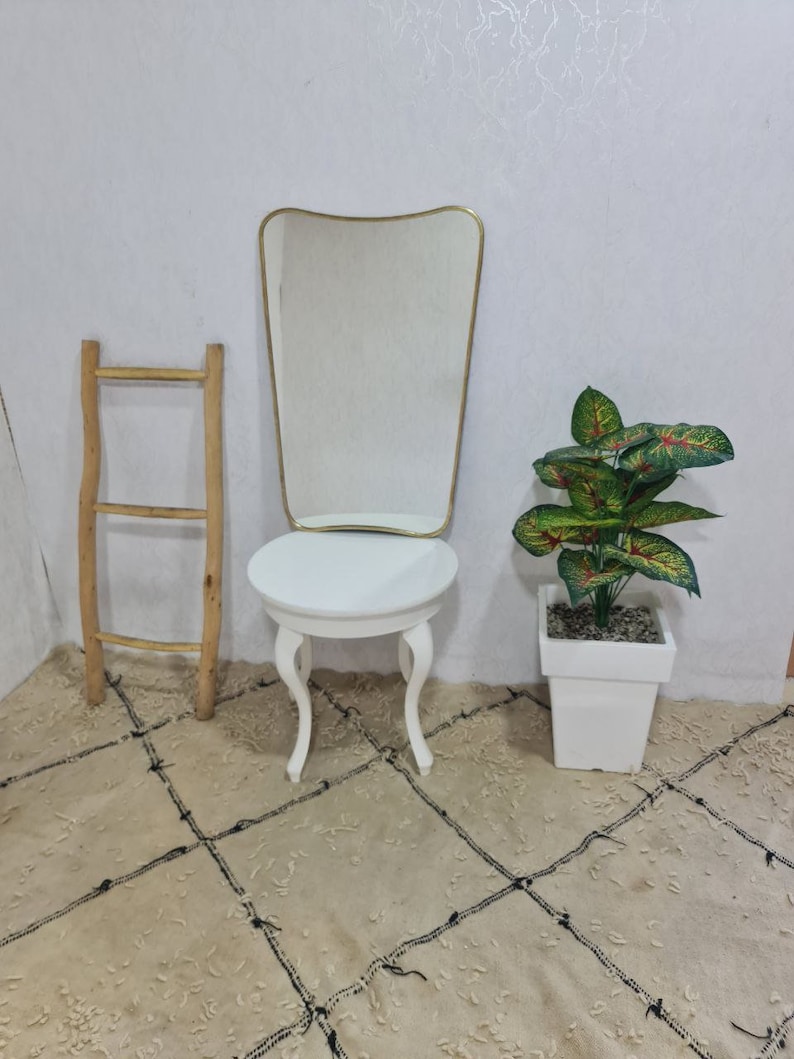May include: A white vanity table with a round top and three legs. A gold framed mirror with a wavy top sits on the table. A wooden ladder leans against the wall behind the table. A potted green plant with red leaves sits on the floor next to the table.