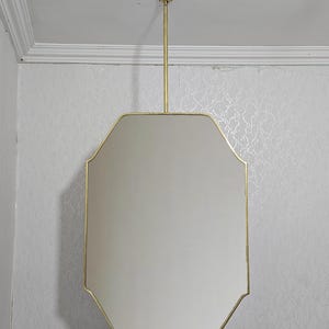 Pode incluir: Um espelho retangular com uma moldura dourada e uma haste de suspensão a condizer. O espelho tem uma forma geométrica única com uma parte superior e inferior planas e laterais curvas. O espelho está suspenso do teto.
