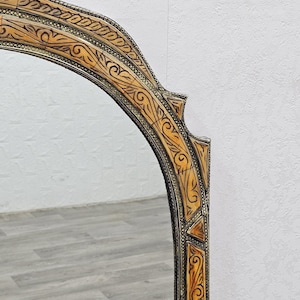 May include: A decorative mirror with an ornate, carved wooden frame. The frame is a light brown color with intricate details and a silver finish. The mirror is oval shaped and has a beveled edge.