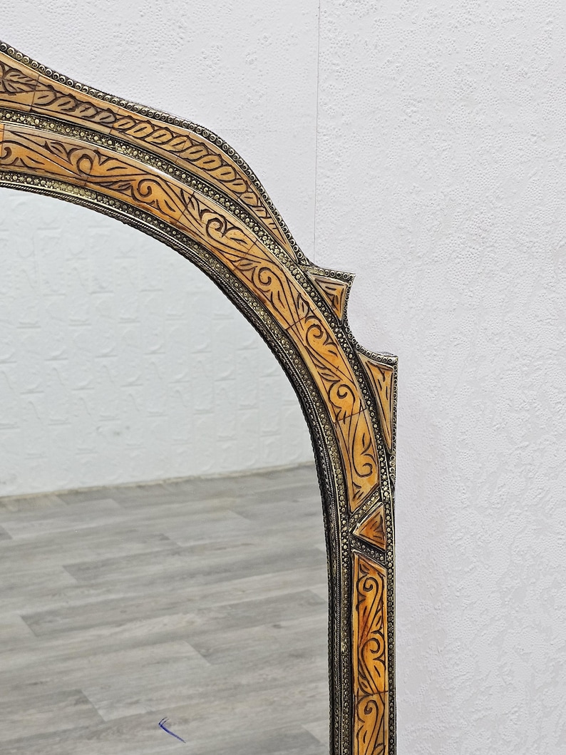 May include: A decorative mirror with an ornate, carved wooden frame. The frame is a light brown color with intricate details and a silver finish. The mirror is oval shaped and has a beveled edge.