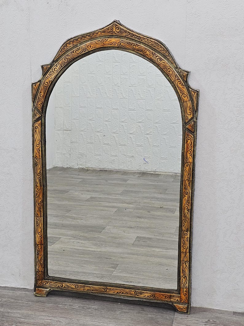 May include: A large, arched mirror with a decorative, ornate frame. The frame is made of wood and is painted in a light brown color. The frame has a carved design with intricate details.