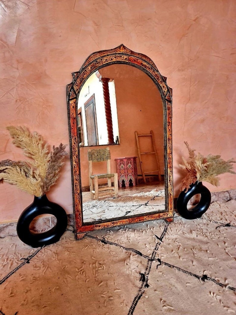 Op de afbeelding: Een grote, sierlijke, gebogen spiegel met een houten lijst. De lijst is geschilderd in tinten oranje, bruin en goud en heeft ingewikkelde houtsnijwerk. De spiegel reflecteert een kamer met een houten stoel en een ladder.
