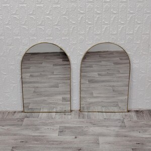 Op de afbeelding: Twee gouden spiegels met een boogvorm en een houten vloer in de reflectie.