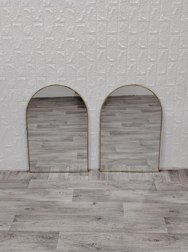 Op de afbeelding: Twee gouden spiegels met een boogvorm en een houten vloer in de reflectie.
