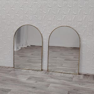 Op de afbeelding: Twee gouden boogspiegels met een witte achtergrond. De spiegels reflecteren de vloer, die van licht hout is.